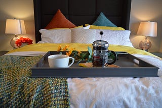 Hotel feel bedroom with bright orange and blue cushions crisp white sheets lamps and tray placed on bed with coffee and flowers