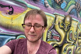 The author is standing in front of a wall covered in colorful graffiti. It’s raining. He has his eyes closed and is not smiling. He has glasses, short brown hair, and a beard. The graffiti includes an alien from outer space. The rest is abstract art.