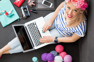 Bird’s Eye view of a blond woman looking up. She is sitting on the ground with a silver laptop on her lap surrounded by a teal folder, a camera, iPad, cell phone, and piles of colorful balls of yarn. From Find Your Niche On Medium by Gema Mora and Canopy Invocations