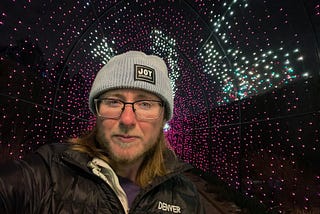 The author is surrounded by white and purple lights. He’s wearing a knitted hat that says “Joy as resistance.” He’s wearing black rimmed glasses and has a beard with enough white to betray that he’s middle-aged. He has long brown hair and a black coat that says “Denver” over a purple sweater. Purple glitter eyeliner matches the lights behind him.