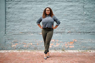 A photo of a confident black woman standing against a blue brick background in a power pose.