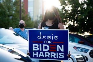 A woman wearing face mask holds a sign that says “desis for Biden Harris.”