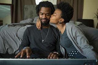 A photo of a black couple. He is playing keyboard and she kisses him on the cheek.