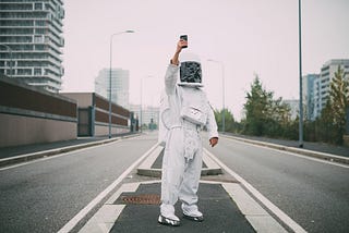 Person wearing astronaut standing in empty street with phone raised, taking a selfie.