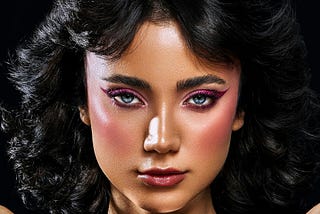 A front-on image of a woman with black hair, blue eyes, and pink eyeshadow/eyeliner. She’s posing with her hands pressed on a disco ball, the backdrop for the image is black.