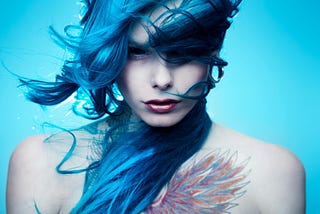 A non-binary person with long, blue hair and black contact lenses stares intensely into the camera lens. They have a colorful tattoo of wings on their chest, and the background is light blue.