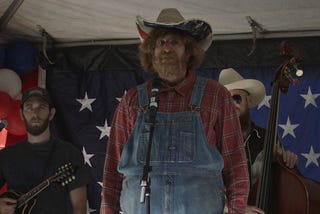 Borat undercover as “Country Steve” on a stage with musicians behind him at a March for Our Rights rally.