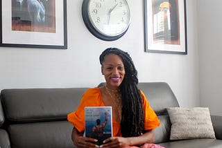 Martinique Lewis sitting on a black couch holding her book on her lap.