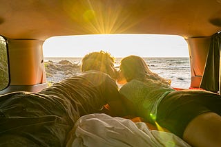 Couple lying down in a van, with the ocean in the background and the sun setting.