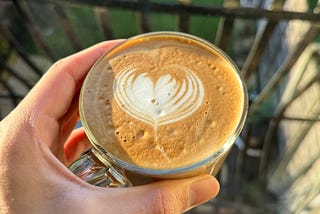 Hand holding a cup of coffee with heart-shape latte art, sun shining, over a garden view from a Juliette balcony.