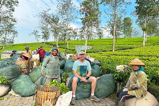 man in tea plantation with workers posing for photos surrounded by bags of harvested tea leaves