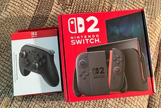 A Switch 2 Pro Controller Box (left) and a Switch 2 Box (right) sitting next to each other on my slightly ugly patterned couch.