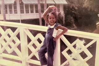 A young girl in blue overalls leans against a white fence. She is doing a ballet pose with one arm curved over her head and the other on her hip. One leg is bent. She’s got a big open-mouth smile.