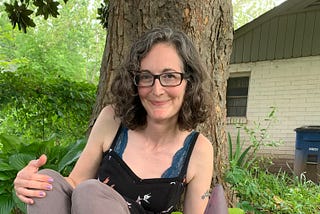 Aimée Brown Gramblin sits under a tree; slender woman with dark brown eyes and dark wavy hair, wearing a lacy cami and a top. Smiles and looks happy under a large-trunked tree. Grass behind.