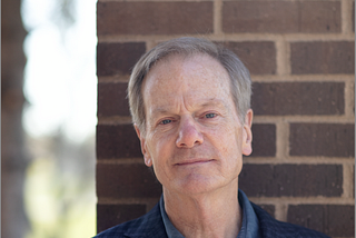Outdoor portrait of William Moyers standing by a brick pillar. He is wearing a blue checked blazer over a denim button-down shirt.
