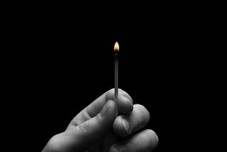 A black and white photo of a person holding up a lit match. The small fire is colored orange.