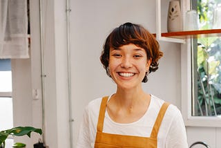 Smiling woman in an orange apron stands behind a wooden counter at what appears to be a bakery. Croissants line the counter. An open window is behind her to the right, a door to the left. White background with plants visible in the room.
