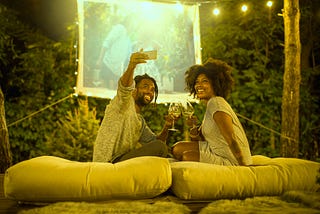 A Black couple take a selfie in their backyard, where they’ve set up a projector screen to watch a movie with hanging lights.