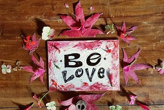 A painting I created on a wooden, worn table of dark stain. It says “Be Love” and is surrounded by leaves I collected