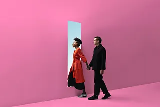 Interracial couple walking through a door in a pink wall, holding hands.