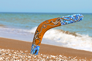 A colorful boomerang, one end of it stuck in the sand on a beach shoreline, symbolizing a rebound, personal growth, healing, reinvention, and life’s unexpected plot twists.