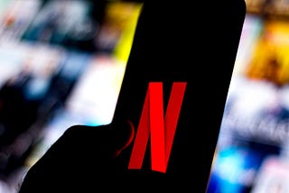 The Netflix logo is shown on a smartphone against a backdrop of blurred out Netflix show titles.