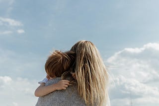 Picture of the back of a person holding a child, set against a blue sky with bright clouds.