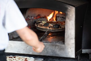 A person using a pizza peel to remove a pizza from an industrial metal wood-fired pizza oven.