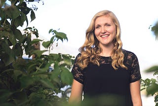 A blond-haired woman in a black shirt standing by a tree and smiling toward the camera