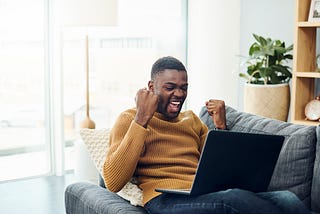 A young man cheers while using a laptop at home on his couch.