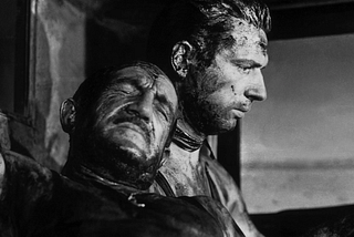 A close up of two men. One man has his eyes closed and is leaning on the other man who is staring down solemnly. Both men are covered in oil and grease. They are in the truck.