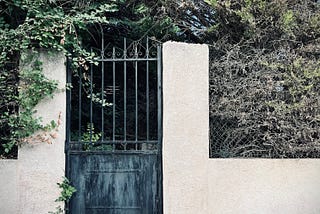 An iron gate in a stucco wall, covered in ivy and surrounded by trees and other overgrowth. The wall is parallel to a steep hill, but it is level, while the ground itself has a considerable slope.