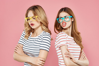 Two young ladies standing back to back wearing big silly sunglasses one looking put off and the other sticking her tongue out with an attitude