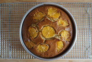 Overhead view of a cake still in its baking tin, resting on a wire rack. The cake is topped with 1 crosswise slice of orange in the center surrounded by 7 other slices.