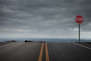 A road that leads off a cliff. A stop sign is right at the edge of the cliff, and the sky is gray and cloudy.