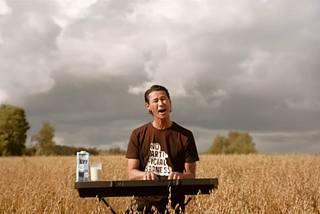 Oatly CEO Toni Petersson playing the keyboard in the middle of a field with a cup and carton of Oatly oat milk to his side.