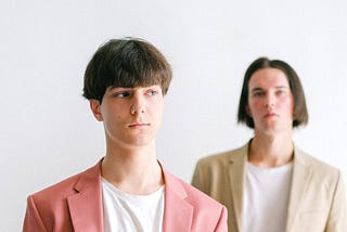 Two men with suit jackets, slacks, and white T-shirts. One has a pink suit and the other has a beige one.