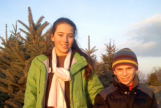 Two kids on a tree farm wearing winter wear; one holds a saw ready to take down a Christmas tree.