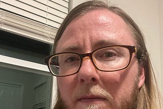 The author is wearing brown framed glasses and a pink shirt. He has a defiant look on his face. He’s standing in front of a window. It’s dark outside.