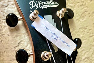 Closeup photo by the female story author of the top neck of a D’Angelico guitar, where a fortune cookie paper fortune strip lays on the strings that reads: “It is never a shame to learn from others.”