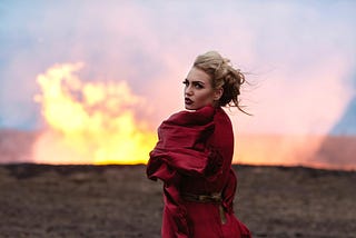 A woman in a red dress, standing in front of a fire looks back at the camera