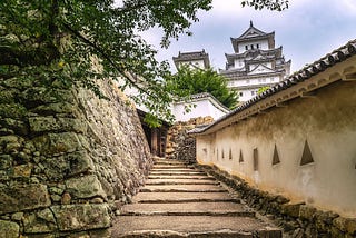 Stone pathway leading up to the main keep of Himeji Castle, flanked by traditional white plaster walls and stone defenses, with the iconic multi-tiered castle visible in the background.