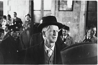 A man wearing an overcoat, vest, tie, and black hat looks off to the side of this black-and-white photograph. There are more men in business formal attire in the background. Over the man’s left shoulder, a man wearing a hat with a badge can be seen looking directly at the camera.