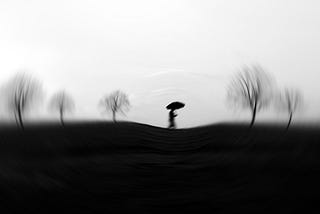 Blurry black and white image of a person walking holding an umbrella with some bare trees.