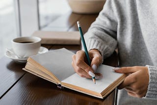 Woman with a cup of coffee, sitting by a window and writing in a blank book.