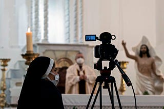 A nun with a video camera on a tripod recording a priest holding the Eucharist at an altar next to a statue of Jesus.