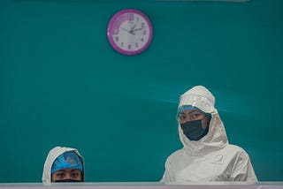 A photo of two healthcare workers wearing masks standing at a counter against a teal background.