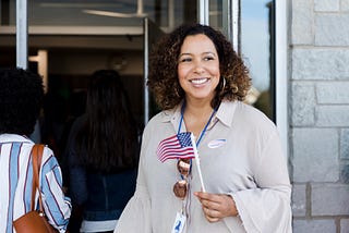 Latina woman holding a little American flag, she has a “Voted” sticker on her shirt.