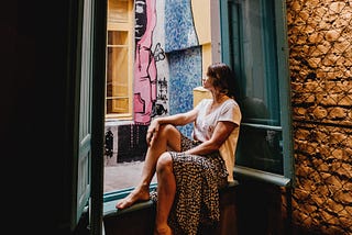 Barefoot woman sits on windowsill, gazing out open window onto a courtyard with street art