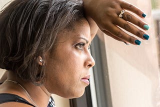 A closeup photo of a black woman leaning against a window and she looks out with a determined expression.